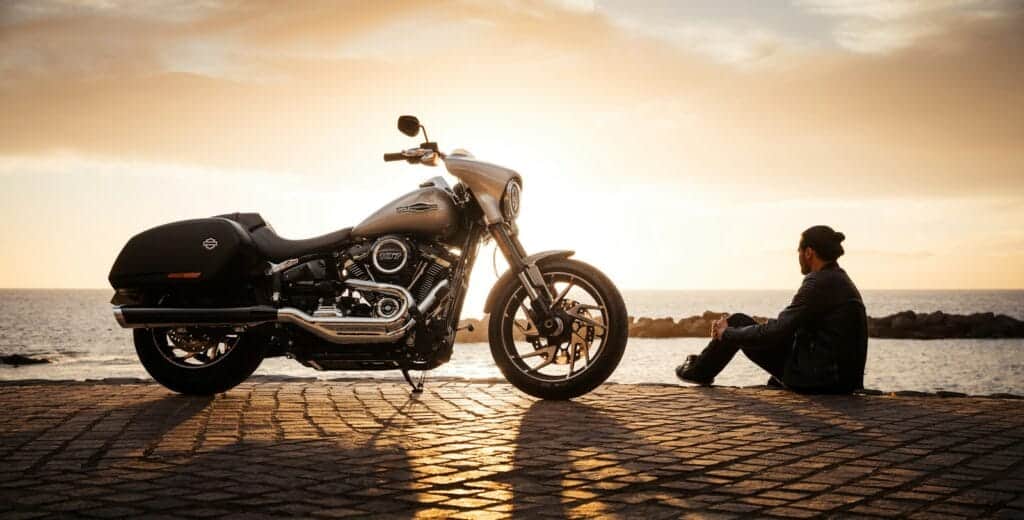 BikesBooking man sitting on ground beside parked silver cruiser motorcycle
