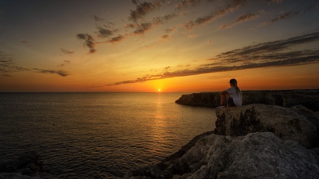 Menorca Travel Guide woman sitting on top of the cliff looking at the horizon of sunrise near body of water
