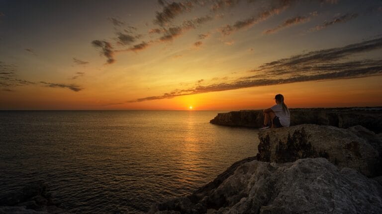 Menorca Travel Guide woman sitting on top of the cliff looking at the horizon of sunrise near body of water