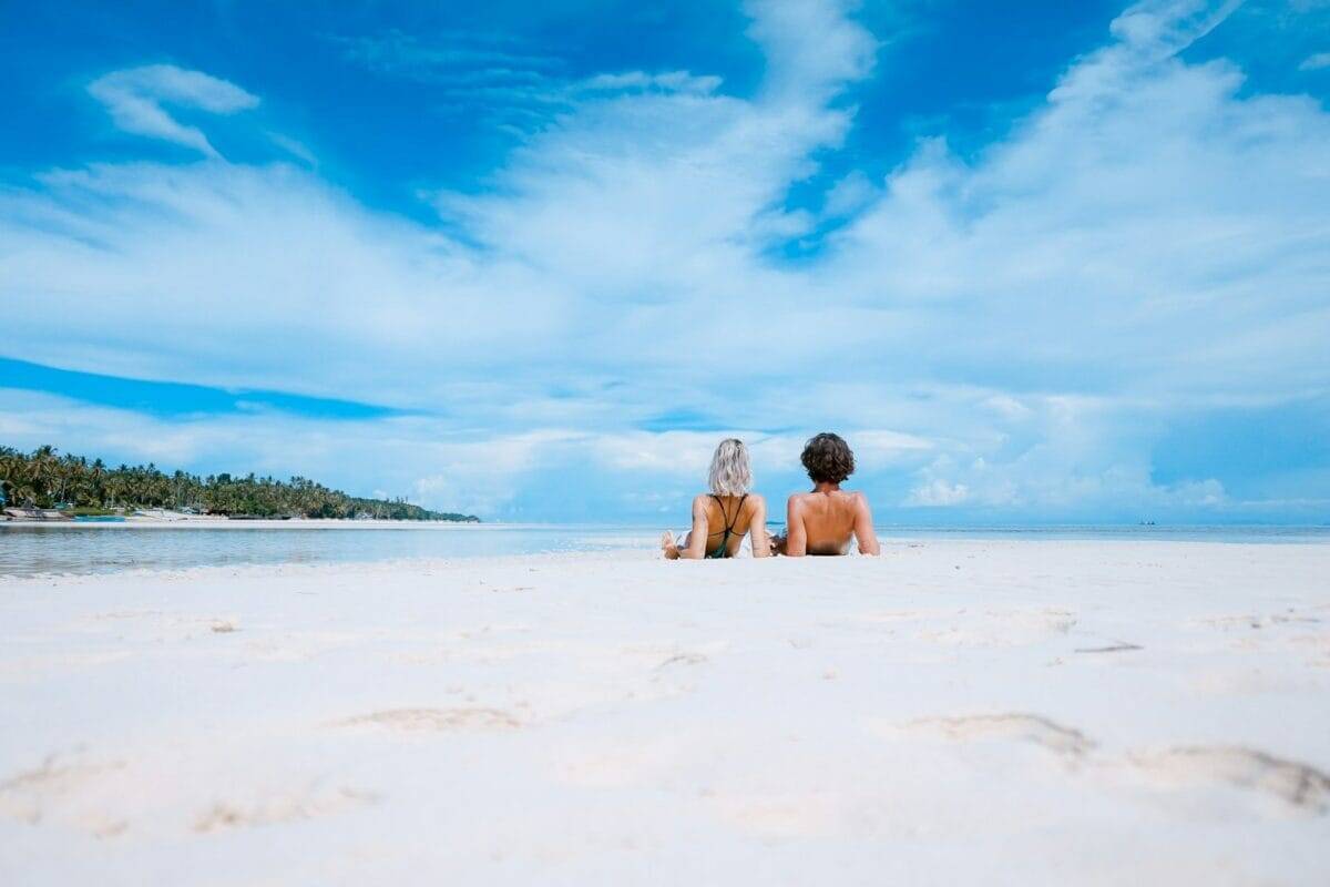 About Us Ideas two women lying on white sand facing beach under blue sky