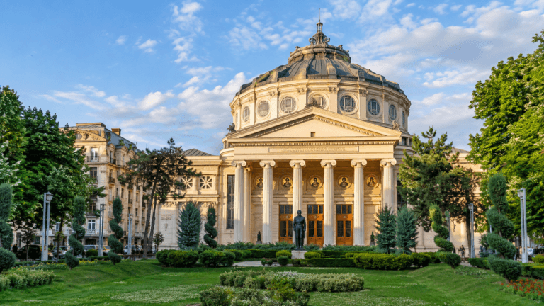 Romanian Athenaeum in Bucharest framed by landscaped gardens and historic architecture beneath a bright blue sky.