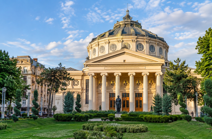 Romanian Athenaeum in Bucharest framed by landscaped gardens and historic architecture beneath a bright blue sky.