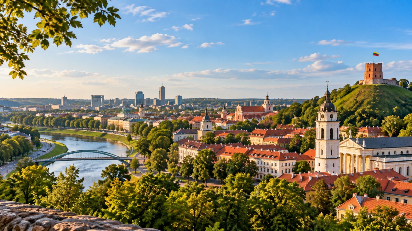 Panoramic view across Vilnius with red-roofed Old Town buildings, the Neris River, church towers, and Gediminas Hill.